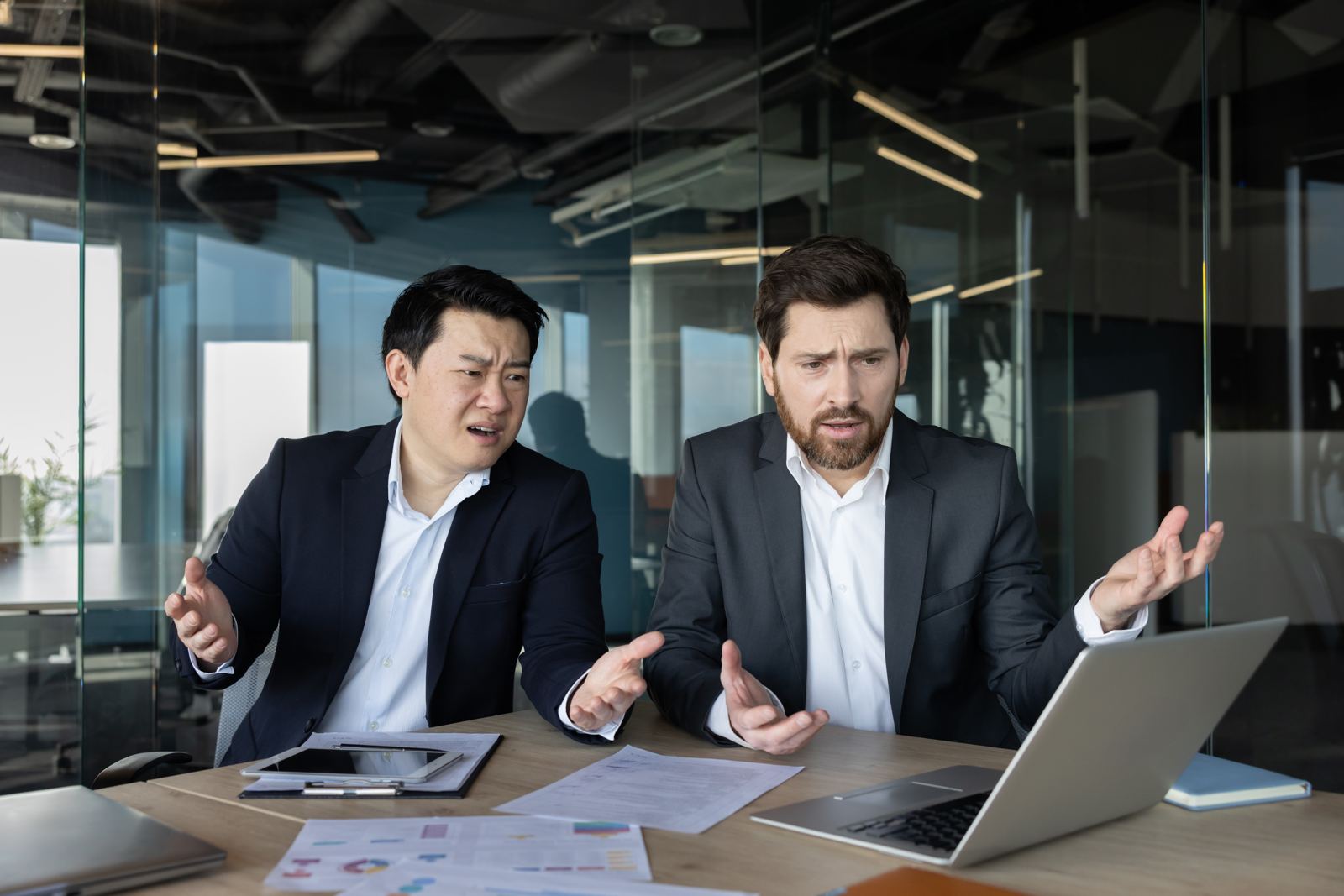 Two businessmen at a desk gesturing in confusion while looking at a laptop
