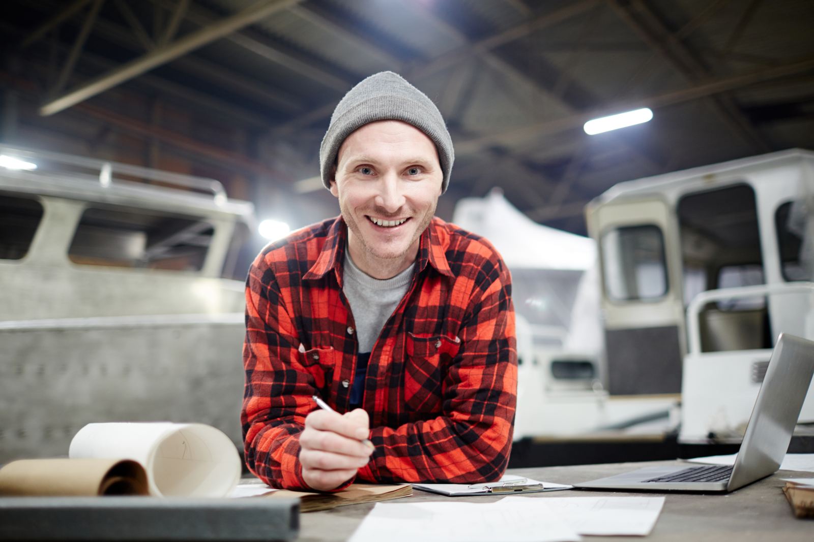 A smiling man in a workshop sitting at a desk with technical drawings and a laptop