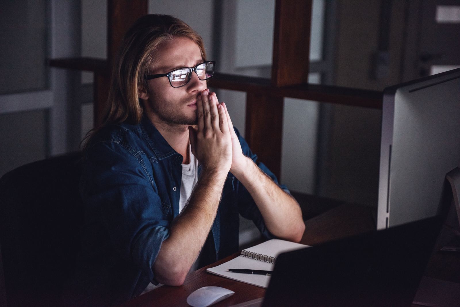 A man sitting at a computer late at night with his hands folded, waiting