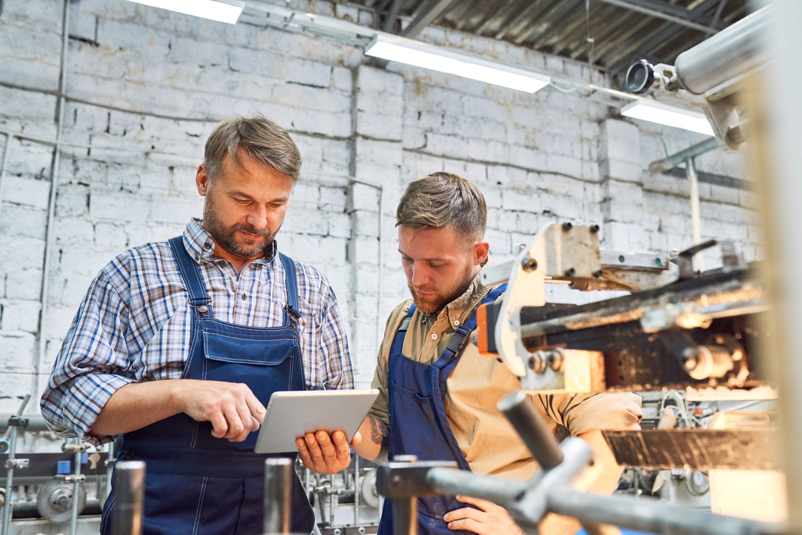 Two factory workers in aprons reviewing information on a tablet next to machinery