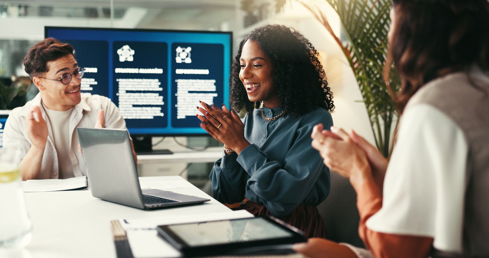 Colleagues at a table applauding together, smiling, with laptops and a screen in the background