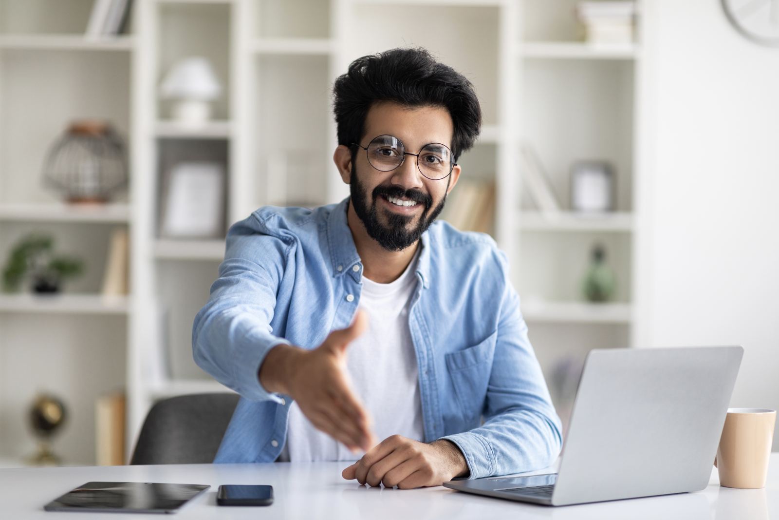 A smiling man at a laptop reaching his hand out towards the camera, as if to shake hands