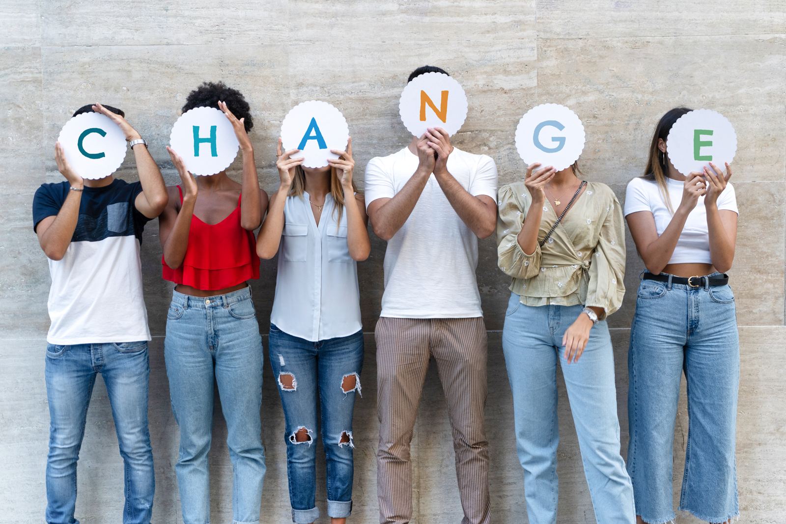 A group of people standing against a wall, each holding a letter in front of their face that together spell CHANGE