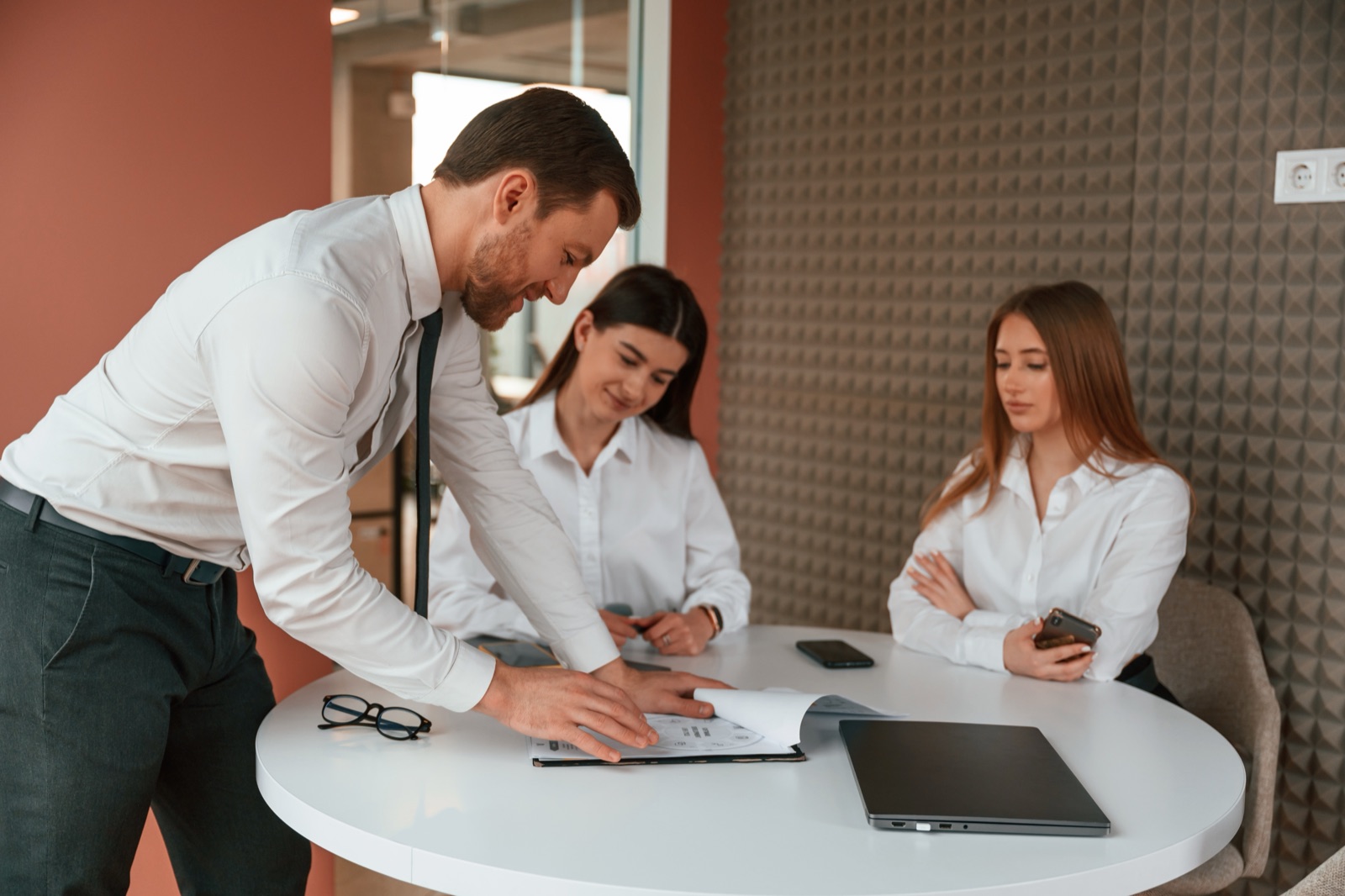 Three colleagues at a round meeting table: a man leaning over documents while two women follow the discussion