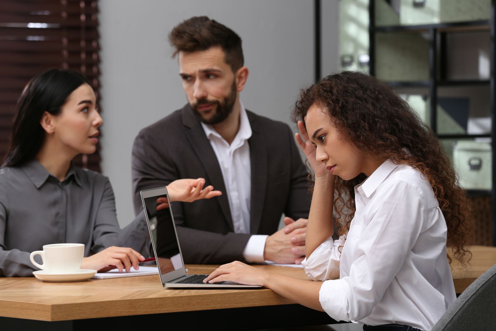 Three colleagues in a tense discussion at an office table, one of them resting her head on her hand
